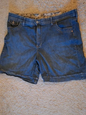 Blå jeansshorts från Lee - Klassiska blå jeansshorts från Lee med fem fickor, knappgylf och bälteshällor. Shortsen har normal passform och är tillverkade i denimmaterial. Bakfickorna har lock och knappdetaljer. Perfekta för varma dagar.