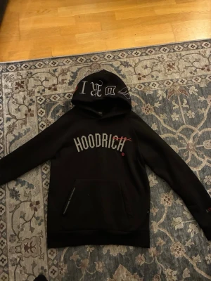 Svart hoodie från Hoodrich med tryck - Svart hoodie från Hoodrich med stort vitt tryck på bröstet och textdetaljer på ryggen. Klassisk huva med tryck och känguruficka framtill. Perfekt för en streetwear-look. Mjuk bomullsblandning för skön känsla.