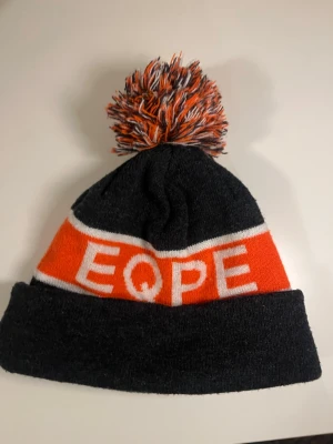 Mörkgrå och orange mössa med tofs - Säljer en stickad mössa i mörkgrått och orange med vit text 'EQPE' runt om. Mössan har en stor, fluffig tofs i matchande färger och uppvikt kant. Perfekt för dig som vill sticka ut med en färgglad accessoar under kalla dagar.