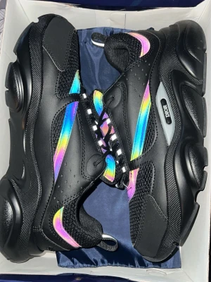 Dior b22  - Svarta Dior sneakers med chunky sula och färgglada regnbågsdetaljer på sidorna och tungan. Skorna har meshpaneler, svarta och vita snören samt reflekterande inslag. Perfekt för dig som vill sticka ut med en unik och modern look.