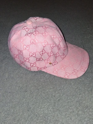 Rosa keps från Gucci med GG-mönster - Snygg rosa keps från Gucci med klassiskt GG-monogram över hela kepsen. Kepsen har böjd skärm och en liten guldfärgad GG-detalj på sidan. Tillverkad i ett slitstarkt tyg med strukturerad känsla. Perfekt för dig som vill sticka ut med en lyxig accessoar.
