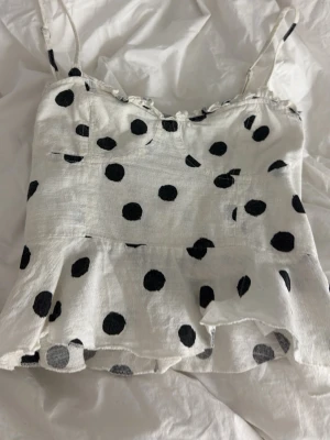 Vit prickig linnetopp med volang - Söt vit linnetopp med svarta prickar och tunna axelband. Toppen har en volang nertill som ger en lekfull känsla. Perfekt för varma dagar och enkel att matcha med jeans eller kjol. Står ingen storlek men gissar på xs