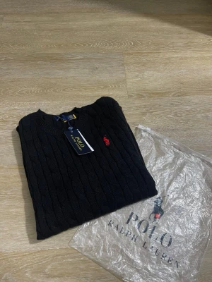 Svart kabelstickad tröja Polo Ralph Lauren - Svart kabelstickad tröja från Polo Ralph Lauren med rund halsringning och röd broderad logga på bröstet. Tröjan har ribbade muddar och är tillverkad i mjukt material. Perfekt för en stilren och klassisk look.