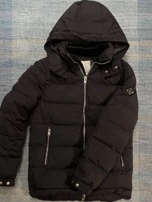 Svart dunjacka med huva och dragkedja - Snygg svart dunjacka med huva och två dragkedjefickor framtill. Jackan har quiltad design, långa ärmar och silverfärgade detaljer vid dragkedjor och knappar. Perfekt för kalla dagar och enkel att matcha med olika outfits.