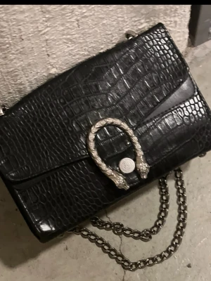 Svart axelväska med kedja och krokopräglat mönster - Snygg svart axelväska i krokopräglat konstläder med silverfärgad kedjerem och dekorativt spänne framtill. Väskan har en rektangulär form och cool metallkedja som axelband. Perfekt för dig som gillar trendiga accessoarer med lite edge.