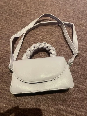 Ljuslila handväska med flätat handtag - Trendig ljuslila handväska i slätt konstläder med ett unikt flätat handtag och justerbar axelrem. Väskan har en stilren, rektangulär form och stängs med lock. Perfekt accessoar för att lyfta din outfit. Lite men så söt💕