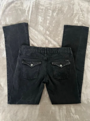 Svarta bootcut jeans från Guess - Snygga svarta bootcut jeans från Guess med dekorativa nitar på bakfickorna. Jeansen har klassisk femficksmodell och silverfärgade knappar. Materialet är mjukt och stretchigt för en bekväm passform. W30 / waist 30 / m. Ungefärliga mått (platta): Över midjebandet: 40 cm Rise: 21 cm Ytterbenslängd: 106 cm Innerbenslängd: 83 cm
