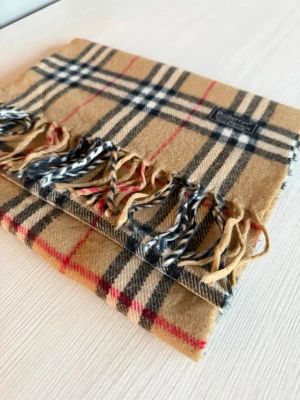 Beige rutig halsduk från Burberry - Klassisk beige halsduk från Burberry i ull med ikoniskt rutmönster i svart, vitt och rött. Halsduken har fransar i ändarna och en vävd logotypetikett. Perfekt accessoar för att lyfta din outfit.