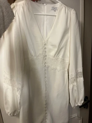 Studentklänning slutsåld från bubbleroom - Elegant vit klänning från By Malina med v-ringning, knappar framtill och spetsdetaljer vid midja och ärmar. Långärmad modell med lätt puff vid ärmslut. Perfekt för dig som gillar romantisk och stilren design.