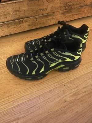 Nike Air Max Plus svarta/gula sneakers - Nike Air Max Plus sneakers i svart med gula detaljer och ikoniska vågformade linjer. Skorna har en sportig siluett, synlig Air Max-dämpning i sulan och mesh-överdel för bra andningsförmåga. Perfekta för dig som gillar streetwear och vill sticka ut.