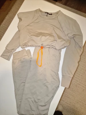 Beige loungewear - set  - Mysigt loungewear-set i beige från Boohoo. Setet består av en långärmad croppad tröja med orange dragsko i midjan och matchande byxor med resår i midjan. Perfekt för chill dagar hemma. Mjukt material och avslappnad passform. Tröjan är I storlek S och byxorna M.