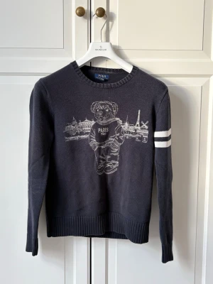 Mörkblå stickad tröja Polo Bear - Mörkblå stickad tröja från Polo Ralph Lauren med Polo Bear och Paris-motiv framtill. Tröjan har två vita ränder på ena ärmen, ribbade muddar och rund hals. Perfekt för dig som gillar streetstyle och ikoniska prints.