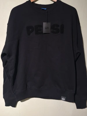 Svart sweatshirt från Pepsi - Cool svart sweatshirt från Pepsi med upphöjd text på bröstet. Tröjan har rund halsringning, ribbade muddar vid ärmar och nederkant samt en relaxed passform. Perfekt för dig som gillar streetwear och vill ha en enkel men snygg look.