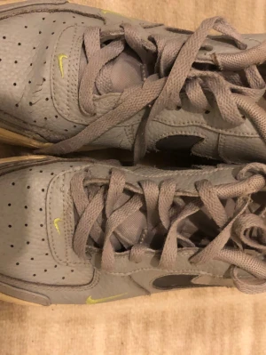 Grå Nike Air Force 1 sneakers - Snygga grå Nike Air Force 1 sneakers med svart swoosh och små gula detaljer. Skorna har klassisk låg siluett, perforerad tåbox och är tillverkade i läder med textilfoder. Perfekta för en streetwear-look. Den är inte tvättad här och skadorna syns ej när man har skorna på sig. Med en tvätt blir de som nya