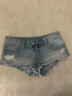 Ljusblå slitna jeansshorts - Säljer ett par ljusblå jeansshorts med slitna detaljer och fransig kant. Shortsen har fem fickor, bälteshällor och knappgylf. Perfekta för varma dagar och en avslappnad stil.