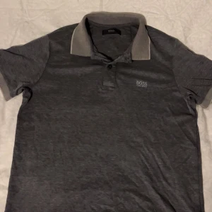 Grå pikétröja från Hugo Boss - Snygg grå pikétröja från Hugo Boss med klassisk krage i ljusare grå ton och knappar framtill. Diskret Boss-logga broderad på bröstet. Perfekt till jeans eller chinos för en clean look.