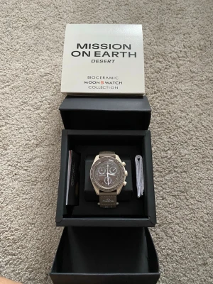 Omega x Swatch MoonSwatch Desert - Stilren beige Omega x Swatch MoonSwatch Mission On Earth Desert från Bioceramic-kollektionen. Klockan har brun urtavla med vita detaljer, tachymeter och tre subdials. Snyggt matchande band med Omega-logga. Perfekt för dig som gillar unik design och samlarobjekt.