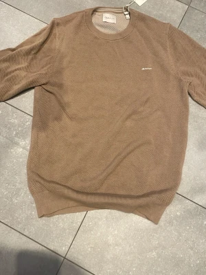 Beige stickad tröja från Gant - Beige stickad tröja från Gant med rund halsringning och diskret logga broderad på bröstet. Tröjan har ribbade muddar vid ärmslut och nederkant. Perfekt för en stilren och avslappnad look.