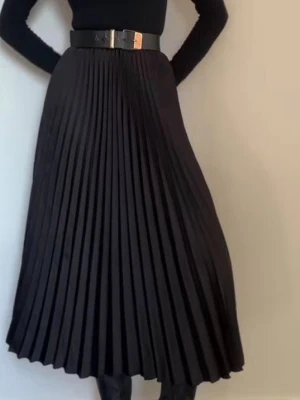 Svart plisserad långkjol - Stilren svart plisserad långkjol med hög midja och markerat midjebälte. Kjolen har ett elegant fall och passar perfekt till både stickade tröjor och blusar. Enkel att matcha och ger en klassisk look.