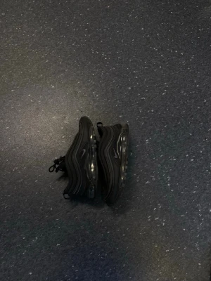 Nike Air Max 97 svarta sneakers - Säljer ett par svarta Nike Air Max 97 sneakers med ikoniska vågiga linjer och reflekterande detaljer. Skorna har synlig Air-sula runt hela foten och är tillverkade i mesh och syntetmaterial. Klassisk Nike Swoosh på sidan och dragflikar både fram och bak.