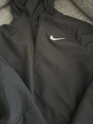 Svart vindjacka från Nike - Säljer en svart vindjacka från Nike då jag har en annan som jag använder så denna tar bara upp utrymme, storleken är 158-170 men jag tycker att den funkar upp till ~175cm. Den är inprincip oanvänd och den köptes för cirka 600 kr. Priset kan diskuteras