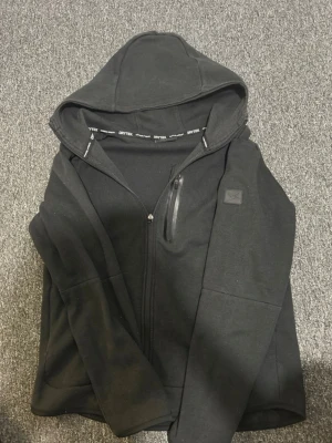 Svart hoodie från Dritek med dragkedja - Säljer en svart hoodie från Dritek med hel dragkedja och huva. Hoodien har en bröstficka med dragkedja och en diskret logga på ärmen. Tillverkad i mjukt och bekvämt material, perfekt för chill eller träning.