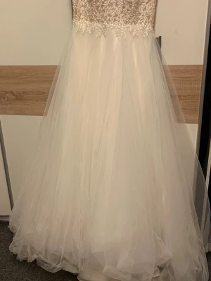 Vit långklänning/bröllopsklänning med spetsdetaljer - Romantisk vit långklänning med vacker spets upptill och voluminös tyllkjol. Klänningen är ärmlös och har fina broderade blommor på överdelen som ger en lyxig känsla. Perfekt för dig som vill sticka ut med en elegant och klassisk look. OBS! Aldrig använd och i original förpackning!!!