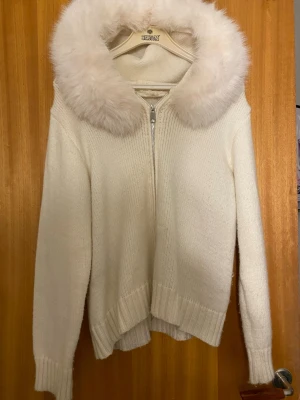 Vit stickad cardigan  med fuskpäls - Mysig vit stickad hoodie med dragkedja framtill och stor huva kantad med fluffig fuskpäls. Snygg och trendig detalj med pälsen runt huvan.