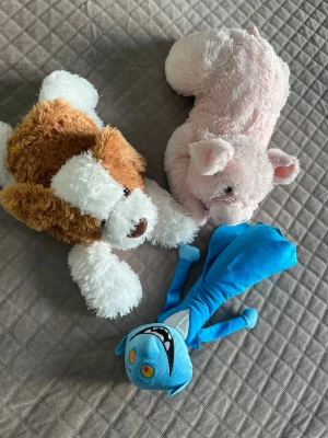 Tre mjukisdjur: hund, gris & blå figur - Säljer tre mjukisdjur: en brun och vit hund, en ljusrosa gris och en blå tecknad figur med stora ögon och vitt leende. Perfekta som gosedjur eller dekoration. Alla är mjuka och gjorda i plyschmaterial.