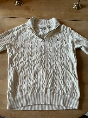 Vit kabelstickad tröja från Gant - Kabelstickad vit tröja från Gant med halv dragkedja och hög krage. Tröjan har ribbade muddar och nederkant samt en liten broderad logga på bröstet. Perfekt för en klassisk och stilren look. 170 i storlek xs isch