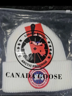 Vit ribbstickad mössa Canada Goose - Vit ribbstickad mössa från Canada Goose med klassisk rund logotyp framtill i rött, blått och vitt. Mössan har uppvikt kant och är tillverkad i ett mjukt, värmande material. Perfekt för kalla dagar och snygg till vinterlooken.