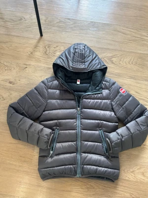 Dunjacka med huva från Colmar - Snygg dunjacka från Colmar med huva och två dragkedjeförsedda fickor framtill. Jackan har quiltad design och Colmar-logga på ärmen och ryggen. Perfekt för kalla vinterdagar och ger en schysstare look om man vill hålla stilen. Använt den ett tag men är för liten. skicket bra storlek 40 i Colmar, Passar nog mellan 155-165 En vinröd/brun färg