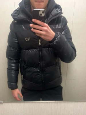 Alessandro zavetti jacka - Säljer en svart pufferjacka från zavetti med glansig finish och ikonisk trekantslogga på bröstet. Jackan har hög krage, dragkedja framtill och två sidofickor med dragkedja. Perfekt för kalla vinterdagar och riktigt snygg streetstil. Helt ny knappt använd säljer för den ej används