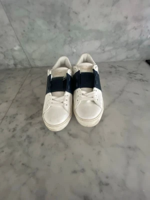 Valentino vita sneakers med blå detalj - Snygga vita sneakers från Valentino med bred mörkblå rem över vristen och klassiska vita snören. Skorna har en stilren design med rund tå och diskret logga på plösen. Tillverkade i läder för en lyxig känsla och passar perfekt till streetwear eller casual outfits.
