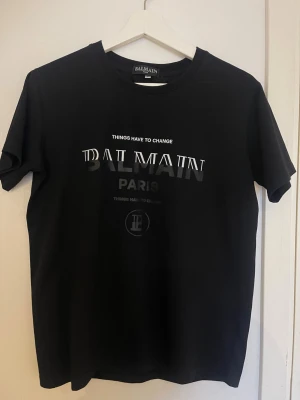 Svart Balmain Paris t-shirt - Snygg svart t-shirt från Balmain Paris med tryck i vitt och grått framtill. Klassisk rund halsringning och korta ärmar. Tillverkad i mjuk bomull för en skön känsla och stilren look. Perfekt för dig som gillar exklusiva streetwear-vibbar.
