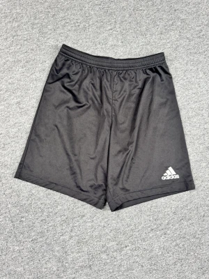Svarta träningsshorts från Adidas - Svarta träningsshorts från Adidas med elastisk midja och klassisk logga på vänster ben. Tillverkade i ett lätt och snabbtorkande syntetmaterial som passar perfekt för sport och aktivitet. Enkel, stilren design utan extra detaljer.