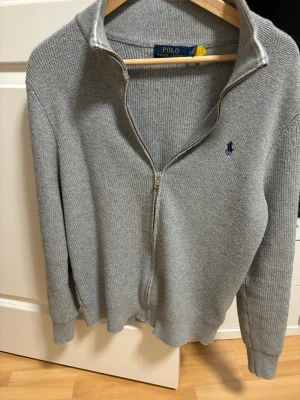 Grå stickad ziptröja från Polo Ralph Lauren - Grå stickad tröja med dragkedja från Polo Ralph Lauren. Tröjan har hög krage, långa ärmar och en broderad marinblå logga på bröstet. Perfekt för lager-på-lager och enkel att matcha med olika outfits.