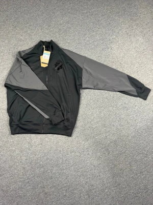 Svart och grå ziptröja AIK - Snygg ziptröja i svart och grått från AIK med klubbens logga broderad på bröstet och stora bokstäver 'AIK' på ryggen. Tröjan har hög krage, hel dragkedja och ribbade muddar. Perfekt för dig som vill representera ditt lag med stil.