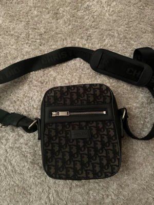 Svart axelväska från Christian Dior - Snygg svart axelväska från Christian Dior med ikoniskt monogrammönster och justerbar axelrem med vadderad detalj. Väskan har en silverfärgad dragkedja framtill och präglad logotyp. Perfekt för dig som vill ha en stilren och trendig accessoar.