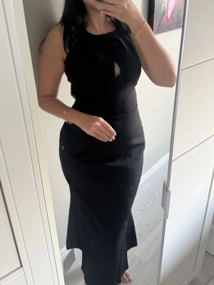 Adoore rozzano midi dress 36 - Elegant svart långklänning med smala axelband och en dekorativ rosdetalj vid halsringningen. Klänningen har en liten öppning framtill och en figurnära passform som framhäver siluetten. Perfekt för dig som vill ha en stilren och sofistikerad look.
