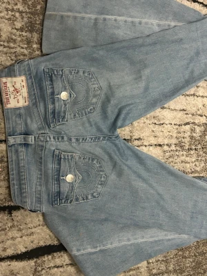 Ljusblå True Religion jeans - Säljer ett par ljusblå jeans från True Religion med klassiska kontrastsömmar och snygga fickdetaljer bak. Jeansen har raka ben och är tillverkade i mjukt denimtyg. Perfekta för dig som gillar en avslappnad men trendig look.