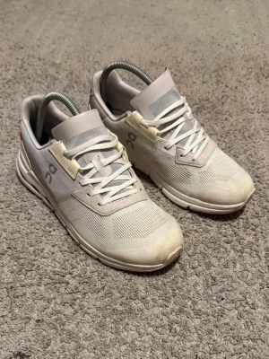 Vita On Running sneakers - Snygga vita sneakers från On Running med mesh-ovandel och klassisk snörning. Skorna har en låg profil och ventilerande material, samt On-logga på sidan. Sulan har On’s karakteristiska CloudTec-dämpning för extra komfort.