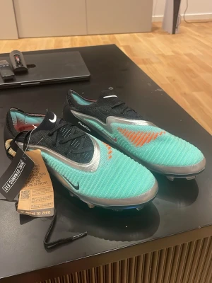 Nike Phantom GT2 turkosa fotbollsskor - Snygga Nike Phantom GT2 fotbollsskor i turkos med svarta och silverfärgade detaljer samt orange mönster på ovansidan. Skorna har låg profil, snörning och ovandel i textilmaterial för bra bollkontroll. Perfekta för dig som vill sticka ut på planen.
