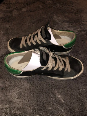 Svarta sneakers med gröna detaljer - Snygga svarta Philippe Model sneakers med gröna detaljer på hälen och vita sulor. Skorna har gråa skosnören och en klassisk låg profil. Tillverkade i läder med märkets logga och texten PM/78 EDT på sidan. Perfekta för en avslappnad streetstyle.