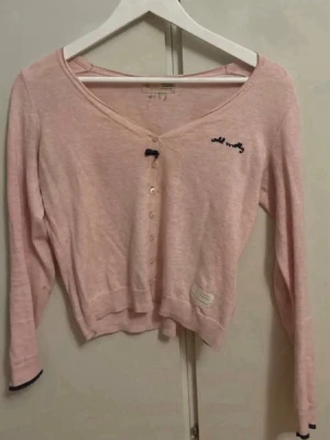 Ljusrosa cardigan från Odd Molly - Söt ljusrosa cardigan från Odd Molly med v-ringning och knappar framtill. Tröjan har långa ärmar, liten rosett vid halsen och broderad logga på bröstet. Tillverkad i mjukt, tunt material och har en avslappnad, croppad passform.
