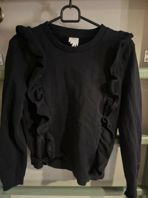 Svart sweatshirt med volangdetaljer - Snygg svart sweatshirt med långa ärmar och volangdetaljer längs framsidan. Rund halsringning och mjukt material som ger en avslappnad men trendig look. Perfekt för dig som gillar en enkel men ändå unik stil.