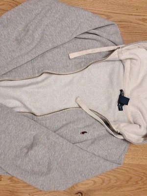 Ljusgrå hoodie från Polo Ralph Lauren - Snygg ljusgrå hoodie med dragkedja från Polo Ralph Lauren. Klassisk design med huva och vita snören, samt det ikoniska röda Polo-märket broderat på bröstet. Mjuk insida och ribbade muddar för extra komfort. Perfekt för en avslappnad stil.