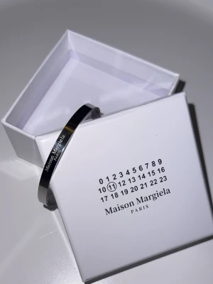 Maison Margiela Silverarmband - Snyggt och stilrent armband från Maison Margiela i silverfärgad metall. Armbandet har en minimalistisk design med ingraverad logotyp på framsidan. Perfekt accessoar för dig som gillar exklusiva detaljer och modern stil. Armbandet är oanvänt (nytt). Kan gå ner i pris vid snabb affär. Skriv vid minsta lilla fundering!