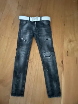 Svarta slitna jeans från Dsquared2 - Säljer ett par svarta jeans från Dsquared2 med slitningar och färgstänk för en edgy look. Jeansen har smal passform och klassisk femficksdesign. Medföljer en vit bälte med stor metallspänne som ger extra attityd till outfiten. Bältet kommer inte med.
