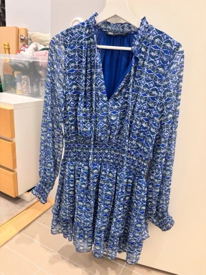 Blå mönstrad kortklänning från Zara - Säljer en blå och vit kortklänning från Zara med långärmad design och elastisk midja. Klänningen har ett blommigt mönster, v-ringning och volangdetaljer vid halsen. Tillverkad i ett lätt och luftigt material, perfekt för varmare dagar.💗